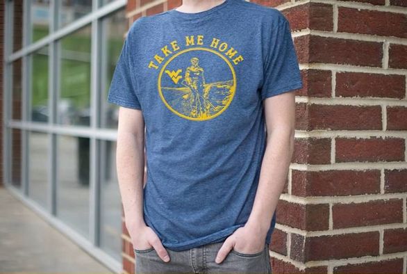 Student wearing the winning shirt. The winning shirt is blue with a gold emblem on the front containing the Mountaineer and the Flying WV. Around the emblem are the words "Take Me Home".