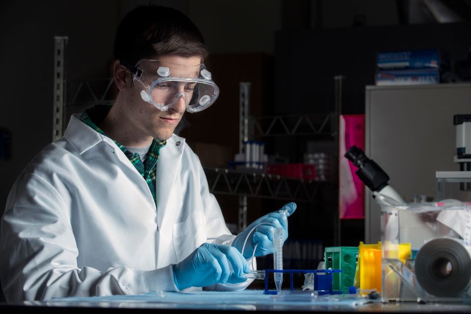 A student with a white coat and gloves works with chemicals in a lab